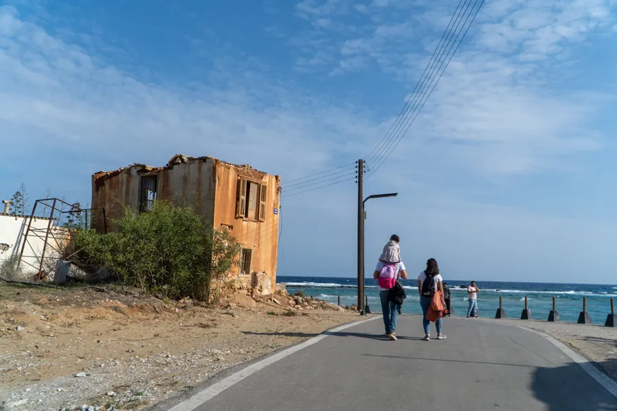 A family walks along a seaside road past a ruined house